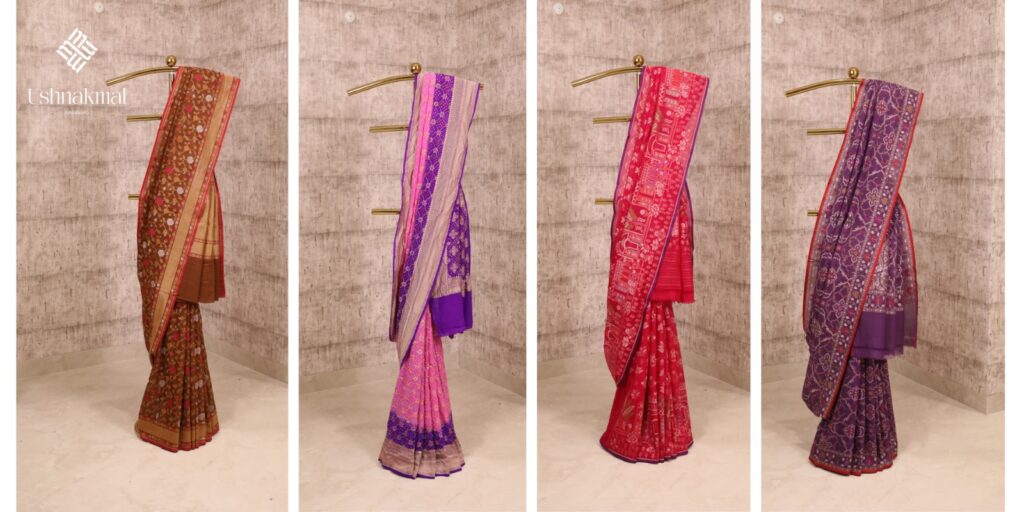 Four designer Banarasi sarees in brown, pink, red, and purple displayed on a gold stand against a beige textured wall.